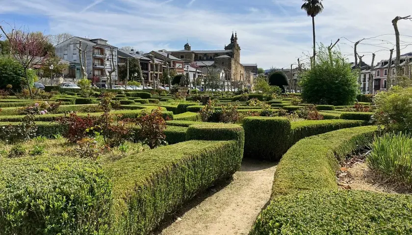 Jardín geométrico con setos cuidados y flores en Villafranca del Bierzo. Al fondo, edificios históricos y una palmera solitaria bajo un cielo azul.