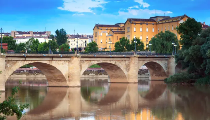 Puente de piedra sobre el río Ebro, reflejándose en sus aguas. Edificios amarillos y verdes árboles componen el paisaje de **que ver en Logroño**.