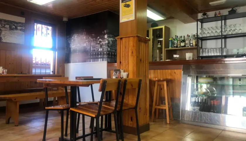 Mesas de madera y sillas listas para comer en Pontedeume. Barra con botellas y vasos, y un mural de la ciudad.