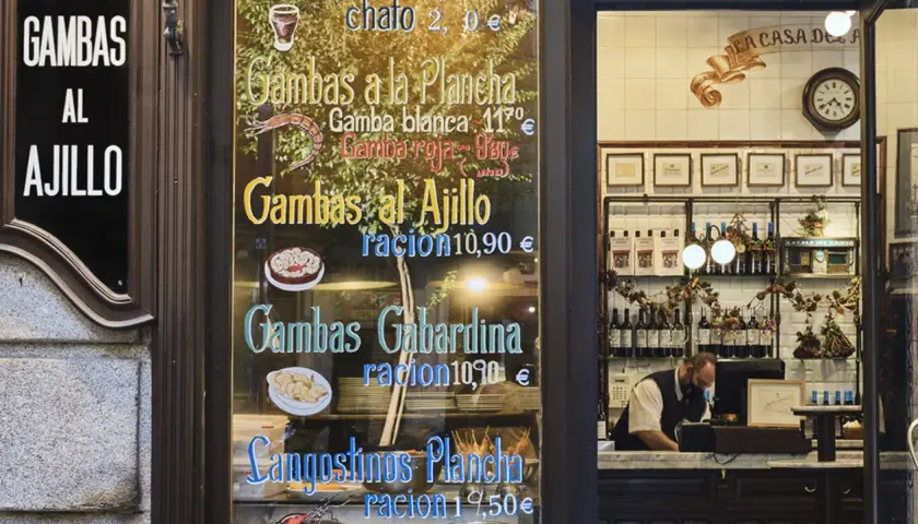 Cartel de un restaurante con menú de gambas y langostinos, y un camarero detrás del mostrador.