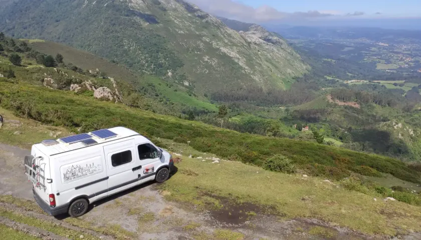 Furgoneta blanca con paneles solares aparcada en una ladera verde, lista para viajar en furgoneta y explorar las montañas.