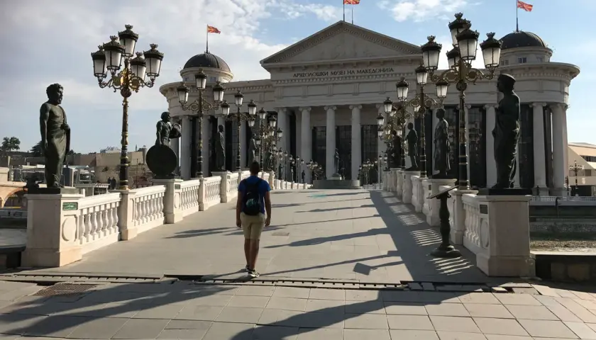 Un hombre camina por un puente adornado con estatuas y farolas, frente al Museo Arqueológico de Macedonia. ¡Un lugar imperdible que ver en Skopje!