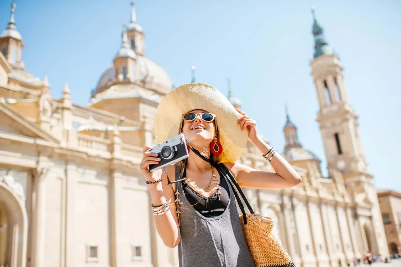 Mujer con sombrero y cámara, lista para viajar por España, con la Basílica del Pilar de fondo.