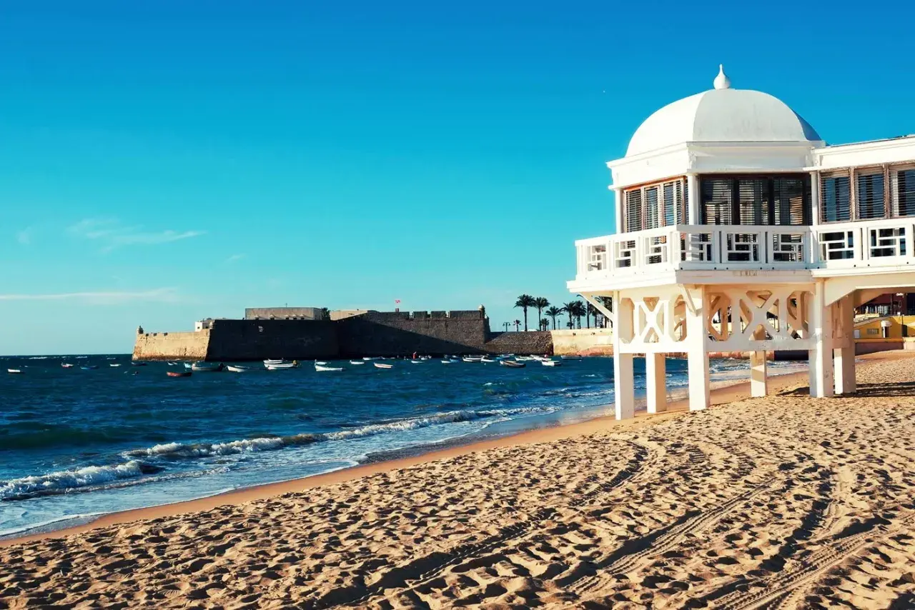Edificio blanco sobre pilotes en una playa de arena, con el mar azul y un fuerte histórico al fondo. Un ejemplo de las mejores playas de Andalucía.