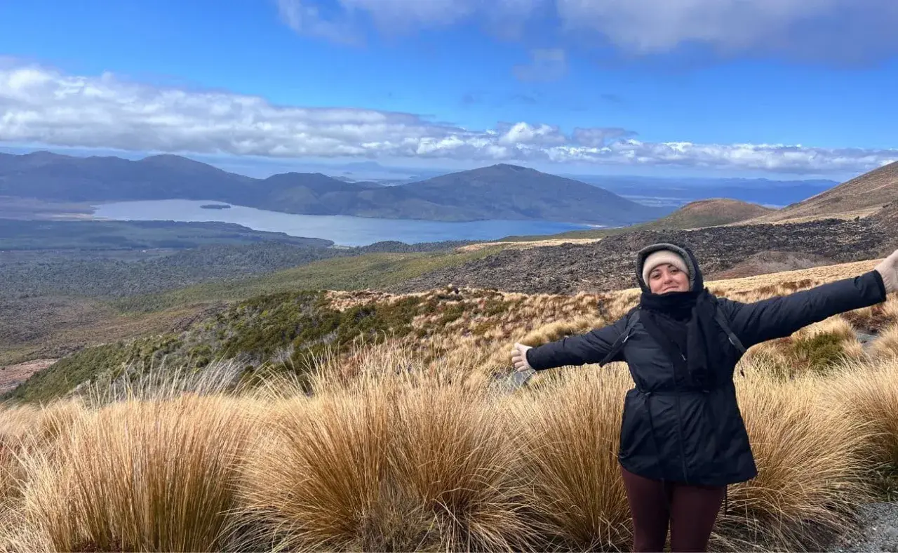 Mujer con los brazos extendidos en el Tongariro Alpine Crossing, disfrutando del paisaje montañoso y el lago azul bajo un cielo nublado.