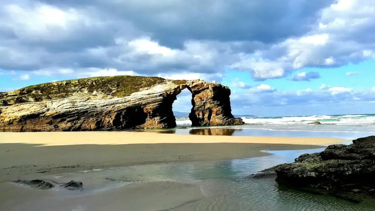 Arco natural en una playa de arena dorada, una de las mejores playas de Galicia, con el mar y el cielo nublado de fondo.