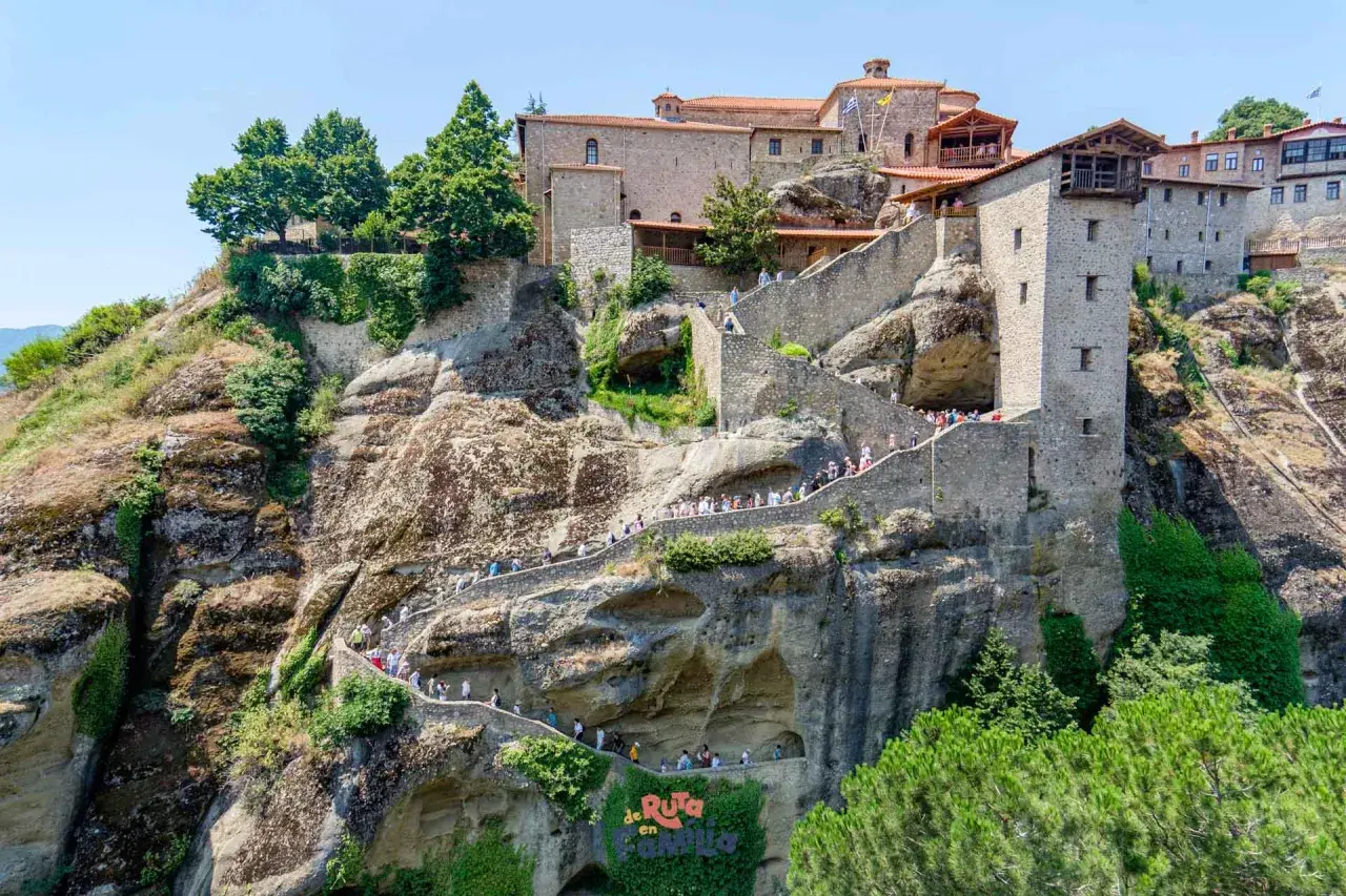 Escaleras de piedra serpentean hacia los impresionantes monasterios de Meteora, construidos en acantilados rocosos. Turistas exploran este sitio histórico.