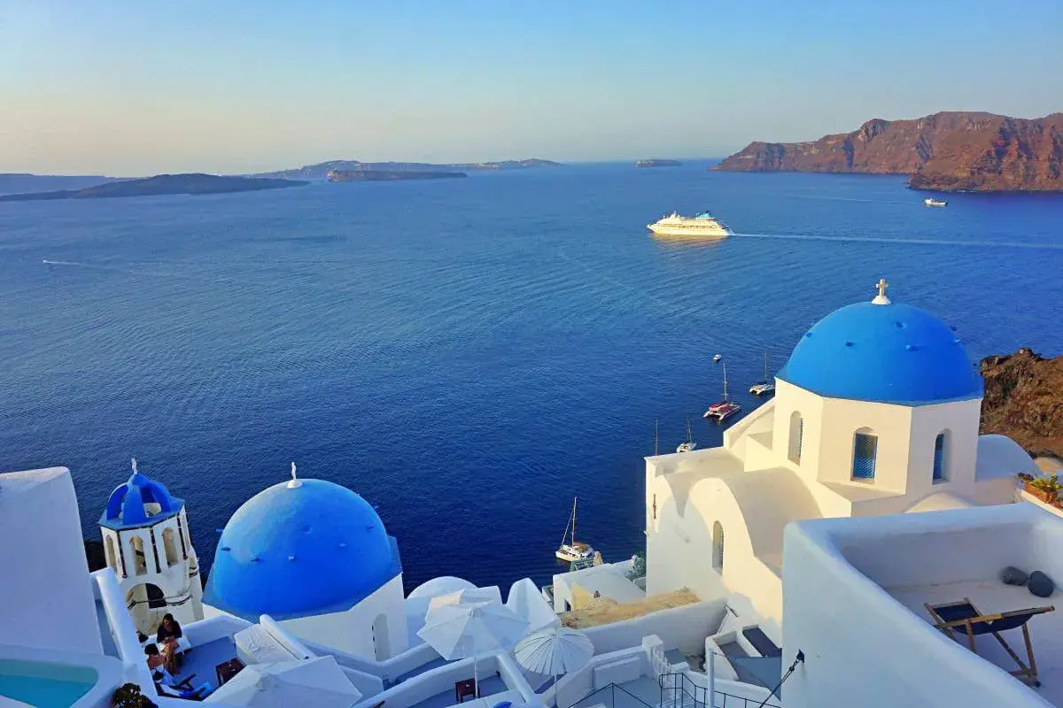 Iglesias blancas con cúpulas azules y el mar Egeo azul profundo. Un crucero navega en la distancia. ¡Imprescindible que ver en Santorini!