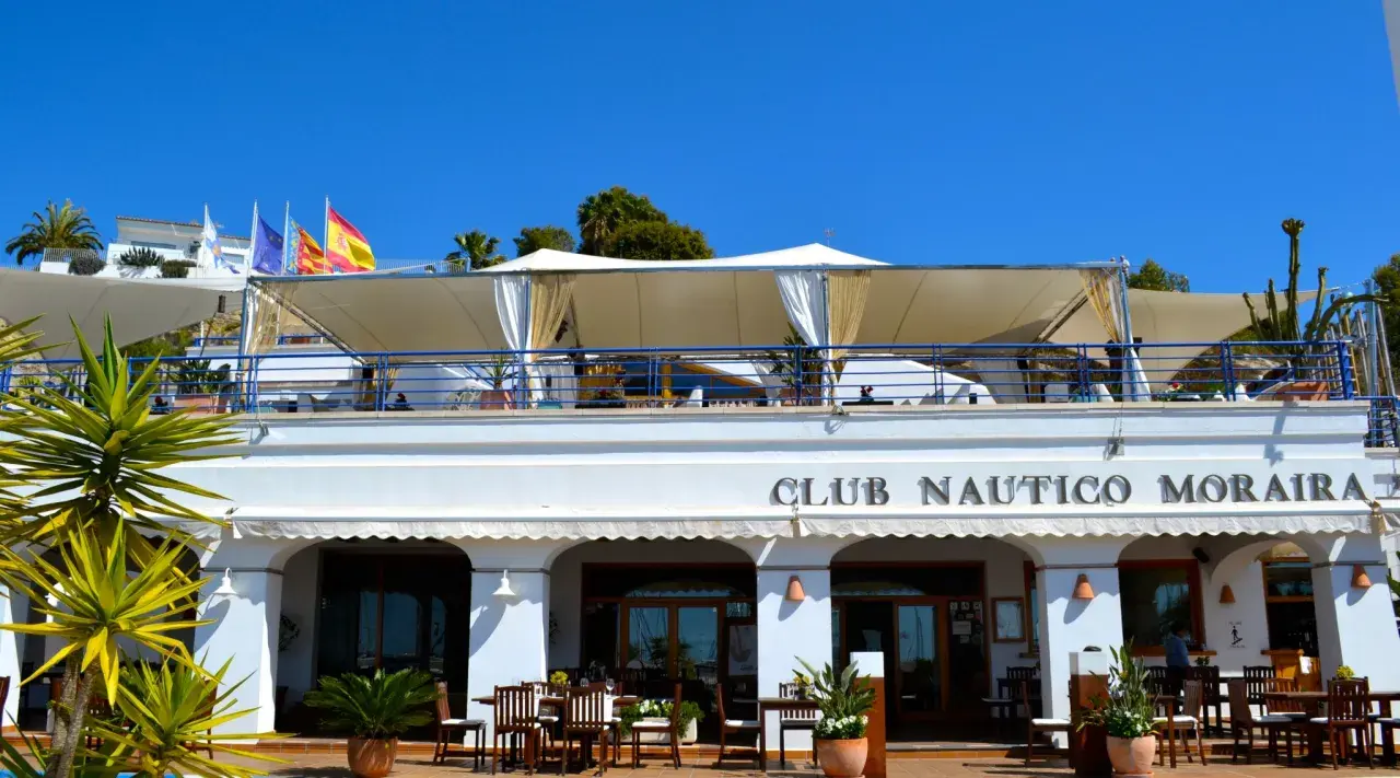 Club Náutico Moraira, ideal para comer en Moraira con vistas al mar. Banderas ondean bajo el sol.