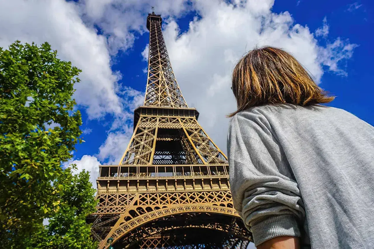 Persona mira la Torre Eiffel, preguntándose cuanto cuesta subir a la torre Eiffel. El cielo azul y las nubes completan la escena.