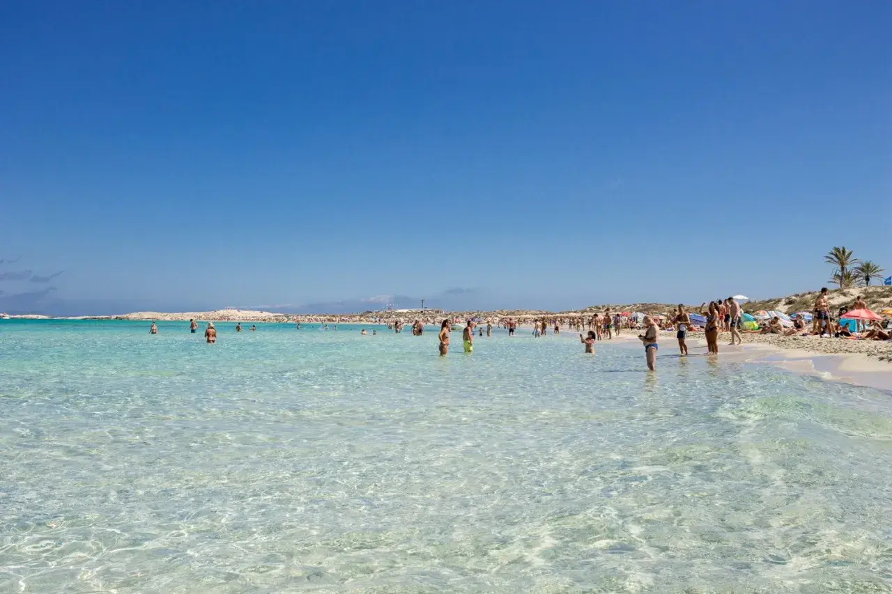 Gente disfrutando de las **mejores playas de Formentera**, con aguas cristalinas y arena blanca bajo un cielo azul.