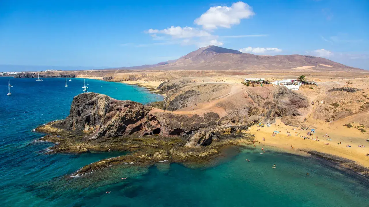 Costa de Lanzarote con playas doradas, aguas turquesas y un restaurante en la colina. ¡Perfecto para tu plan de Lanzarote en 4 días!