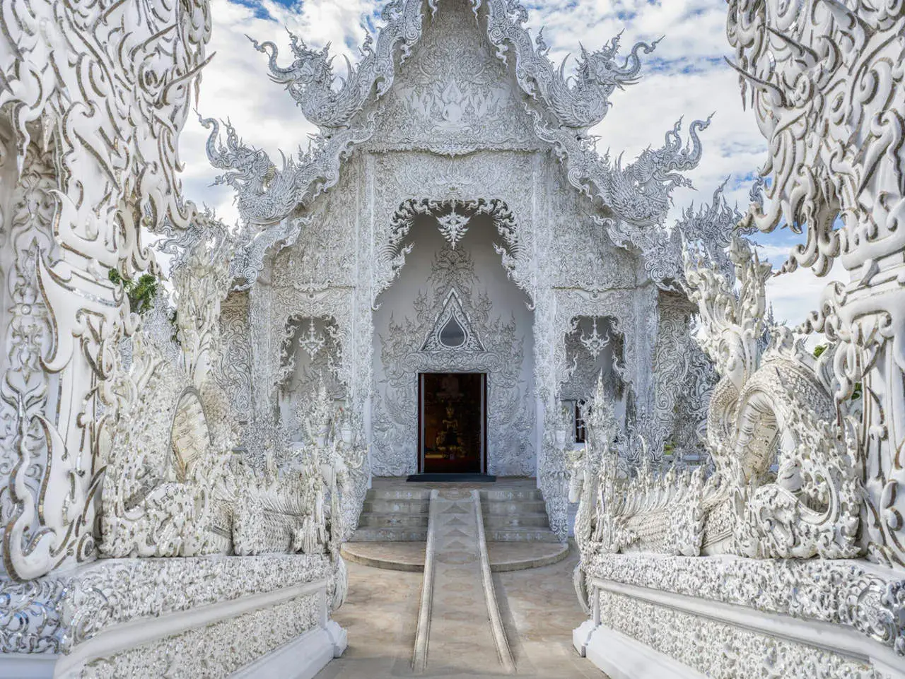 El Wat Rong Khun, un templo blanco deslumbrante en Tailandia, exhibe intrincadas esculturas de dragones y motivos ornamentados bajo un cielo azul.