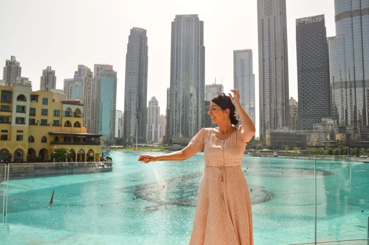 Mujer sonríe mientras disfruta de su viaje a Dubai, con rascacielos y una fuente de agua de fondo.