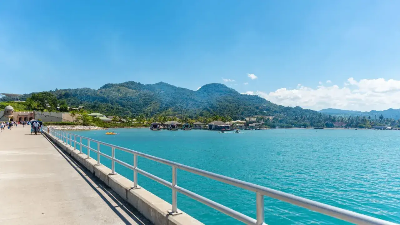 Un muelle con barandilla metálica conduce a un pueblo sobre el agua con montañas verdes de fondo. Ideal para excursiones en Puerto Plata.