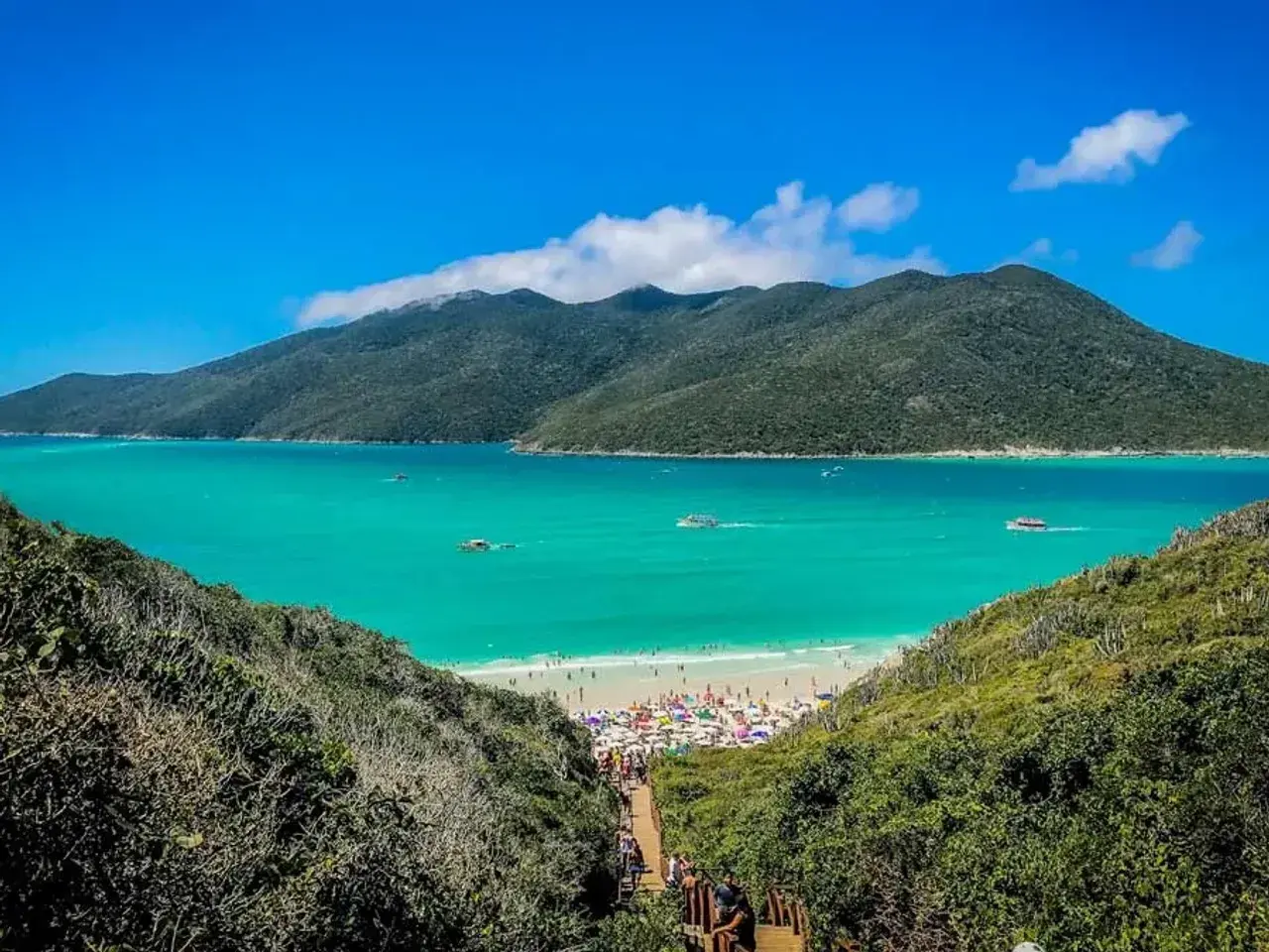 Escenario idílico para viajar a Brasil: playa con aguas turquesas, montañas verdes y gente disfrutando bajo sombrillas.