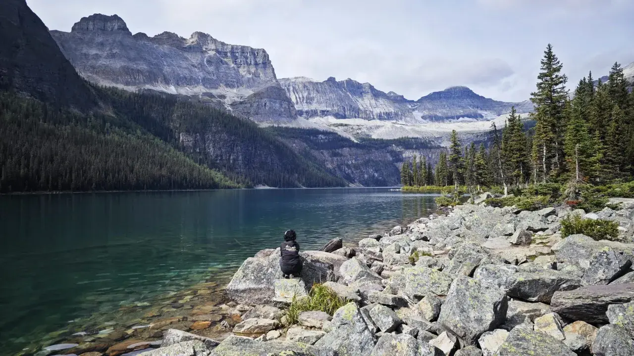 Persona sentada en rocas junto a un lago turquesa, contemplando majestuosas montañas nevadas. Un paisaje perfecto para viajar a Canadá.