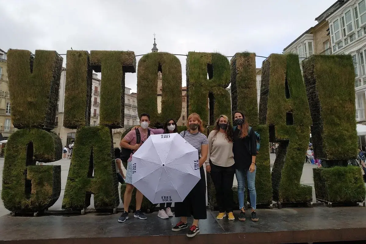 Grupo posa frente a letras gigantes de césped que dicen "VITORIA". Sostienen un paraguas blanco con el logo "FREE TOUR".