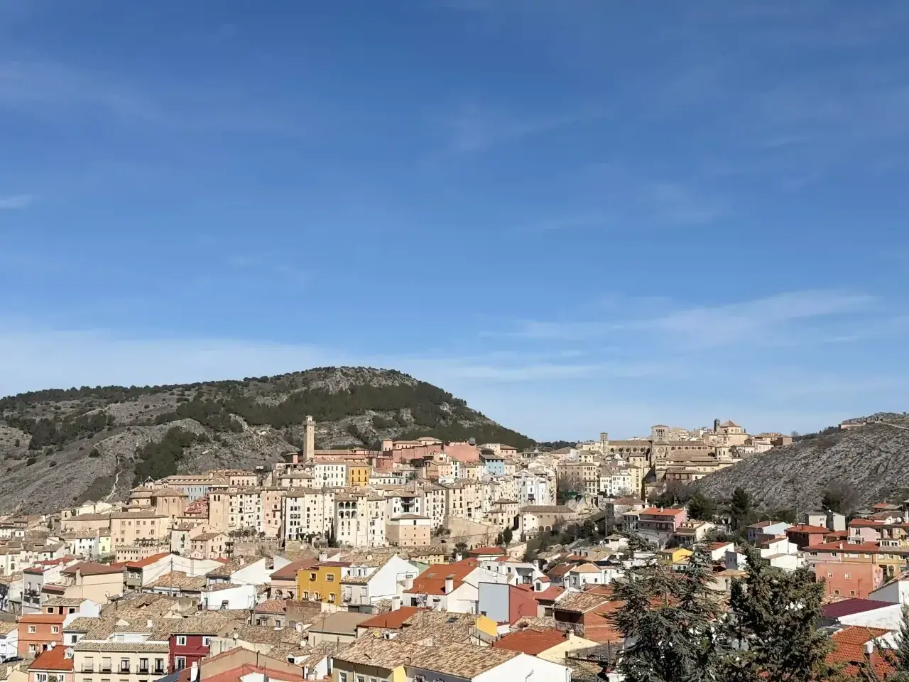 Vista panorámica de Cuenca, con sus casas de colores y tejados anaranjados, asentadas en una ladera rocosa bajo un cielo azul. Ideal para saber qué ver en Cuenca en un día.