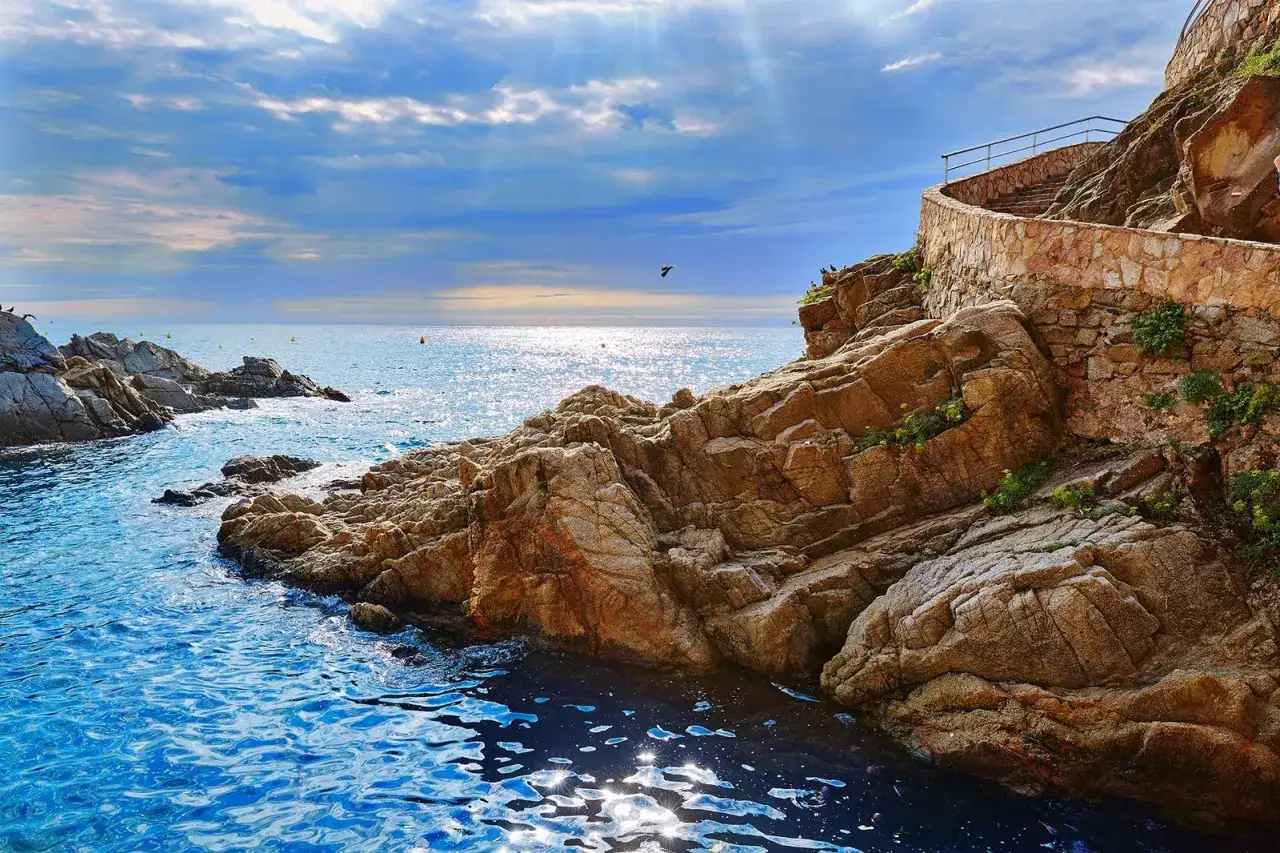 Escaleras de piedra ascienden por un acantilado rocoso junto al mar azul. Un camino de ronda Costa Brava invita a explorar la costa.
