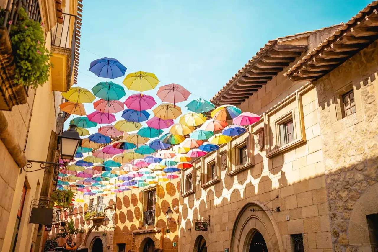Un paseo por calles de Barcelona con paraguas de colores suspendidos, ideal para tu itinerario de qu&eacute; ver en Barcelona en 3 d&iacute;as.