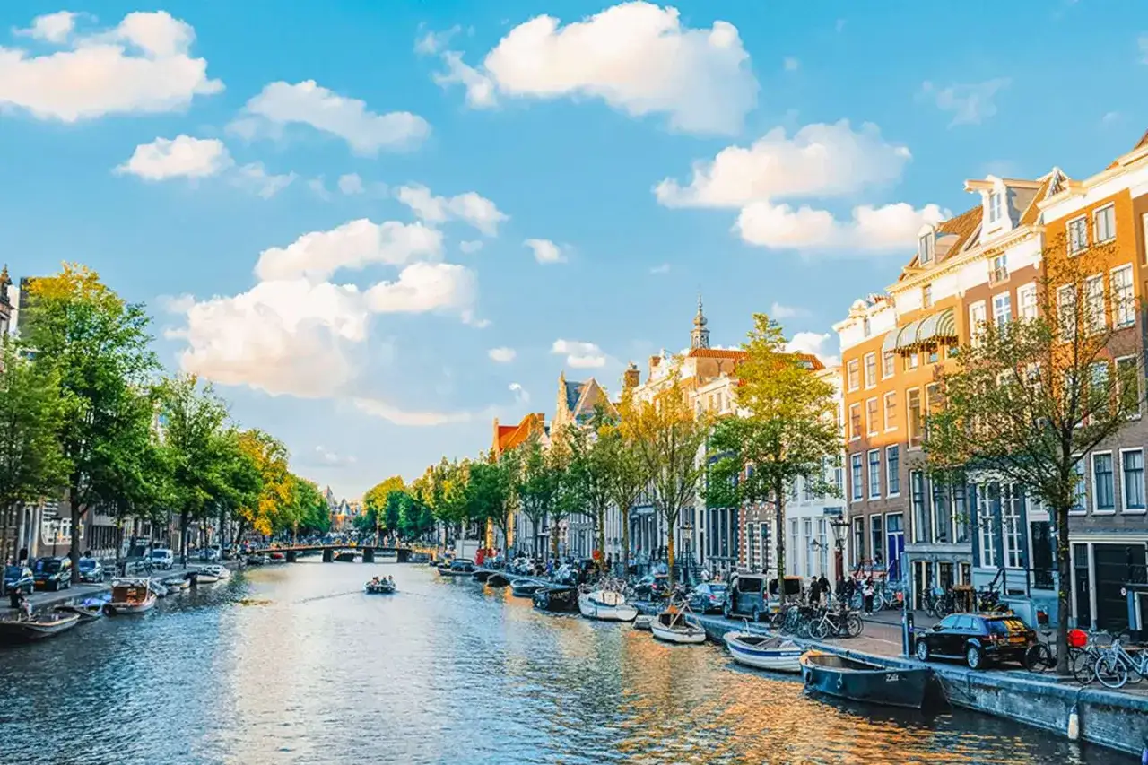 Un paseo en barco por los canales de &Aacute;msterdam es ideal para saber qu&eacute; ver en &Aacute;msterdam en 3 d&iacute;as. Casas hist&oacute;ricas, &aacute;rboles y cielo azul.