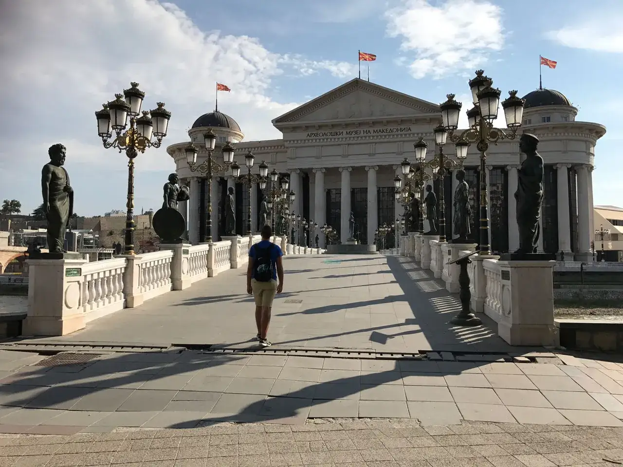 Un hombre camina por el Puente de los Milagros en Skopje, con estatuas y el Museo Arqueológico. ¡Mucho que ver en Skopje!