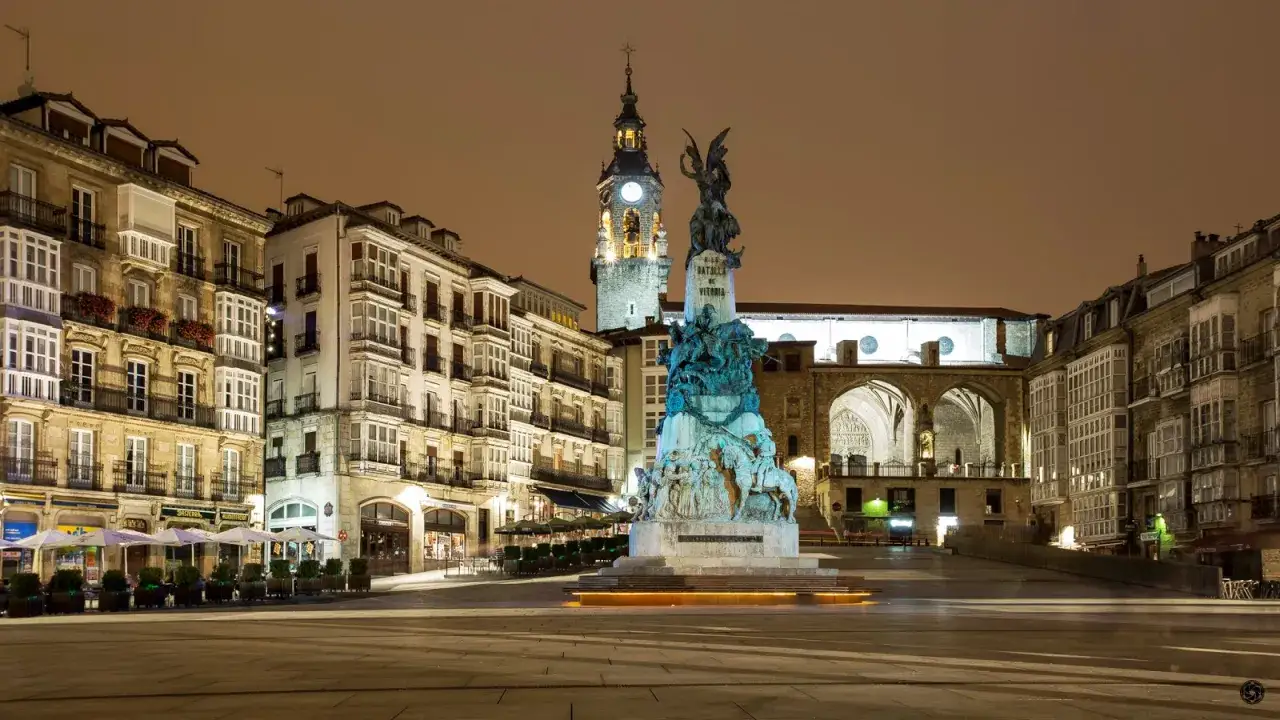 Plaza iluminada de noche con un monumento central. Edificios históricos y una torre con reloj flanquean la plaza, ideal para un free tour Vitoria.