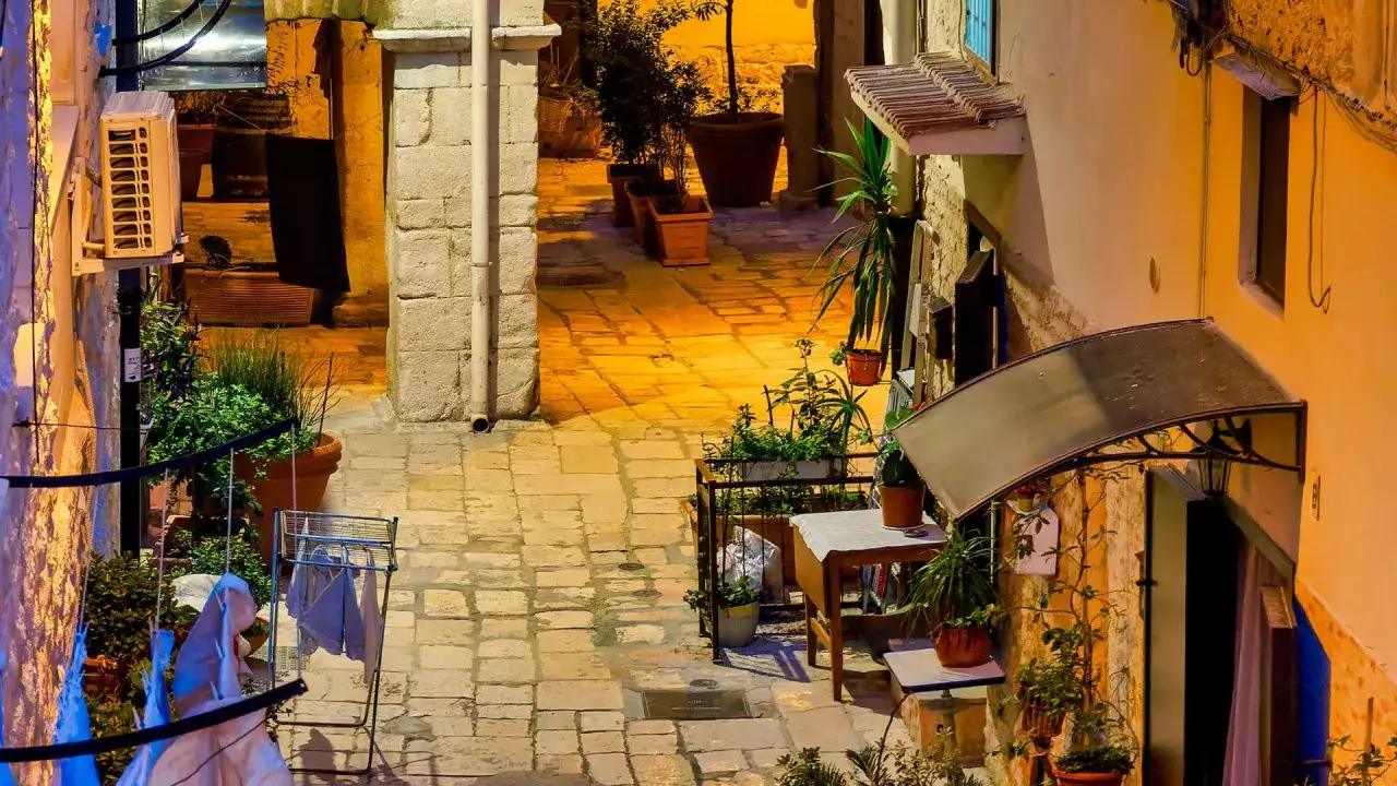 Un callejón con adoquines iluminado por la noche. Plantas en macetas y ropa tendida crean un ambiente acogedor. ¿Qué más bari que ver?