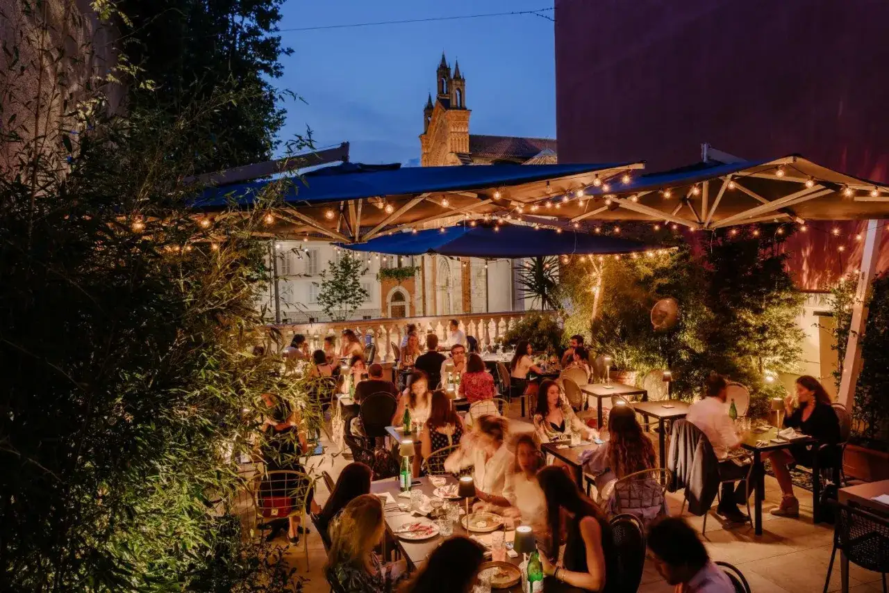 Terraza iluminada con luces de hadas, gente cenando y un edificio histórico al fondo. Un lugar ideal para saber dónde comer en Milán.