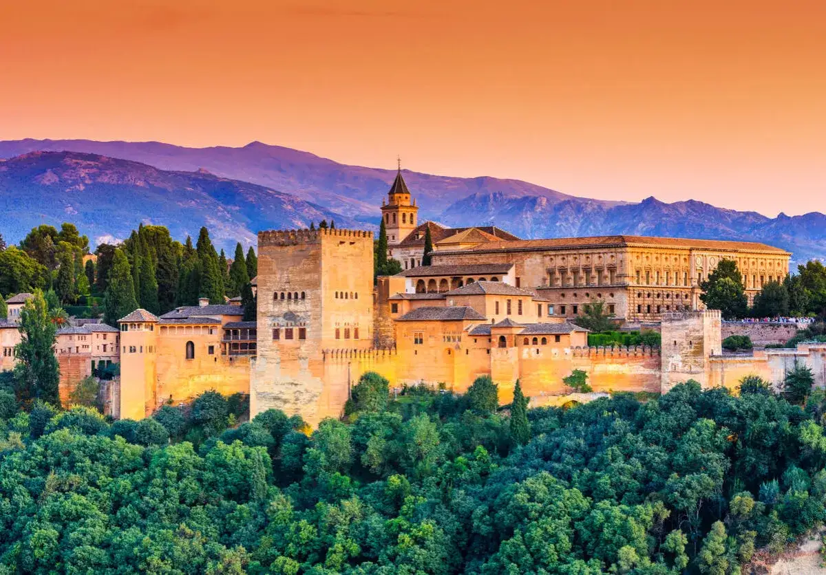 La Alhambra de Granada, un lugar imprescindible que ver en Granada en 3 días, se alza majestuosa entre árboles verdes y montañas al atardecer.