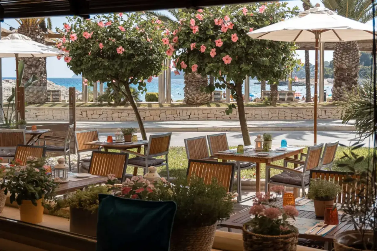 Mesas de restaurante con vistas al mar en Moraira. Flores y árboles en flor adornan el espacio, invitando a comer al aire libre.