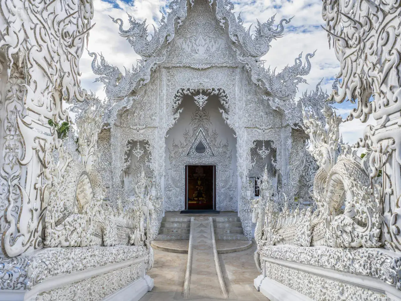 El Wat Rong Khun, un templo blanco deslumbrante en Tailandia, exhibe intrincadas esculturas de dragones y motivos ornamentados bajo un cielo azul.