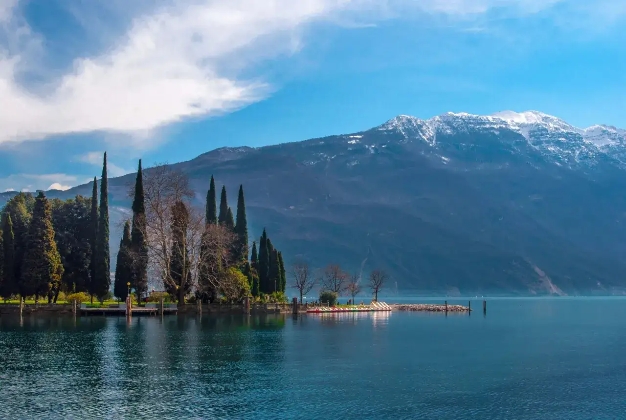 Paisaje sereno en el norte de Italia: lago azul, monta&ntilde;as nevadas y cipreses altos junto a un muelle con barcas de pedales.