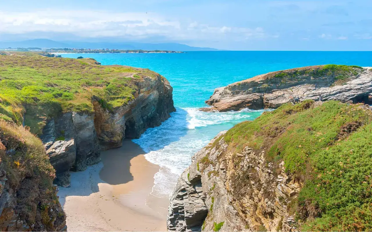 Acantilados verdes y rocosos flanquean una cala de arena dorada y aguas turquesas, un paraíso entre las mejores playas de Galicia.