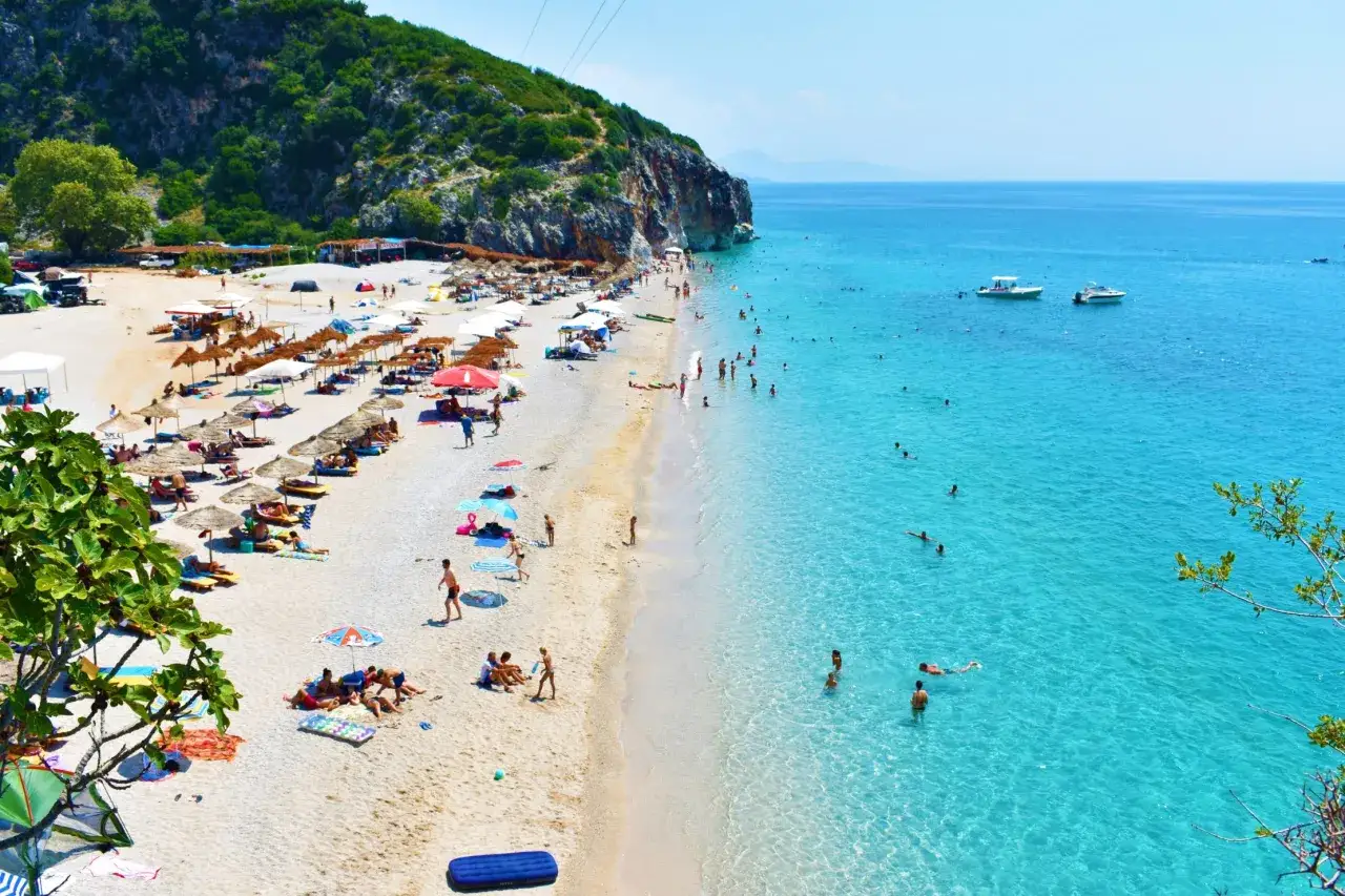 Disfruta de un día soleado en la playa albanesa, con aguas cristalinas y gente relajándose bajo sombrillas. ¡Ideal para viajar a Albania!