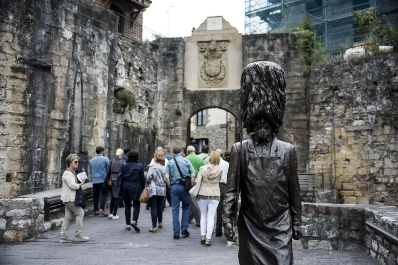 Escultura de hombre con peluca en la entrada de Hondarribia. Turistas pasean por la calle empedrada, descubriendo qué ver en este lugar histórico.