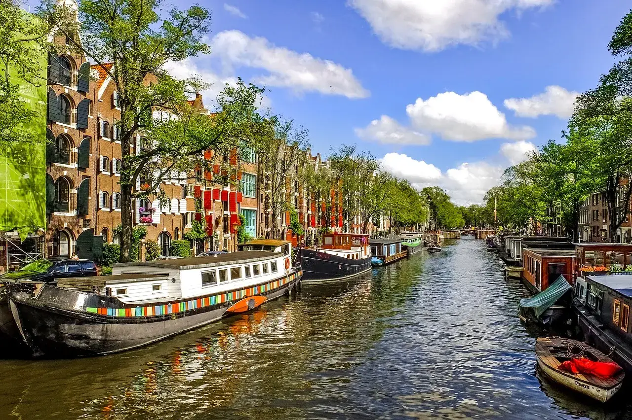 Barcos en un canal de Ámsterdam, un día perfecto para un free tour. Casas con fachadas de ladrillo y árboles frondosos bordean el agua.