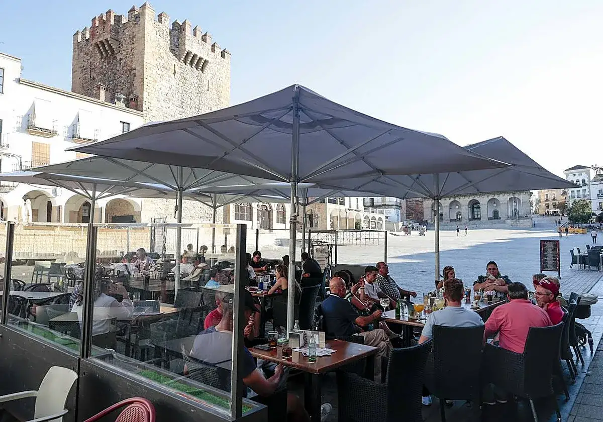 Gente disfrutando en una terraza bajo sombrillas, un lugar ideal donde comer en Cáceres, con la torre de la iglesia de San Mateo al fondo.